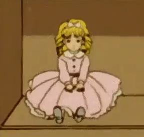 A doll with blonde ringlets, worn in a high ponytail, with a pink bow on top, curling parted fringe, a pink dress with buttons and a darker sash around the waist, frilly white Peter Pan collar, white lacy petticoat showing an extra trim of lace underneath.