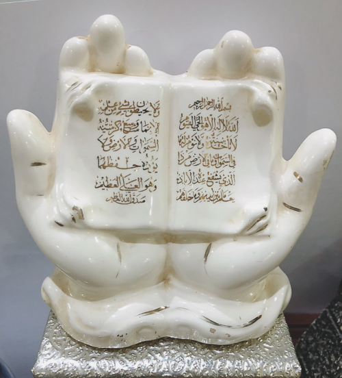 A porcelain figure pair of hands outspread, pressed side-by-side, in a an Islamic supplicating/praying gesture, with Ayat Al-Kursi (Verse of the Throne) written in gold colour across the pages resting in the hands.