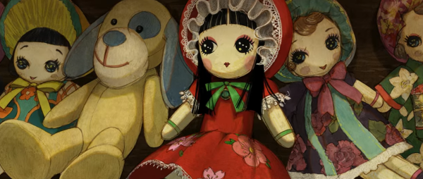 A soft and plush Bunka Doll with straight black hair in a traditional hime cut with blunt fringe, large sparkling black eyes, a line of purple eyeshadow, round face, rosy cheeks, small red lips, pale skin. She wears a red bonnet with white lace, a red dress with short sleeves, also trimmed with white lace. Her European style dress has another line of white lace. She wears a pair of green bracelets on both her short, stubby arms, matching the green of the ribbon tying her bonnet. Her lower half is longer, although her equally stubby feet are out of view. There are other Bunka Dolls around her in different colours and traditional Japanese patterns of cloth, tailored into western fashions.