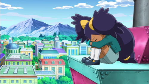 Screencap of Iris, sitting alone with her face buried in her arms, which rest on her drawn-up knees, sitting on a high tower in the city.