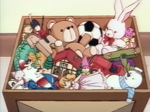 A screencap from Licca-chan no Nichiyoubi, showing a toybox full of toys, including a teddy bear, snowman, fighter plane, rabbit with a red bowtie, soccer ball, toy soldier on a horse, a wooden puppet, another plane, a grey plush cat, a small dollhouse, some sort of jester, and a doll in a pink dress with long wavy orange hair with a straight fringe and a big red bow atop her head, and red shoes.