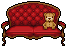 A teddy bear sitting alone on a sofa with red tufted fabric upholstery from Moon Cat.